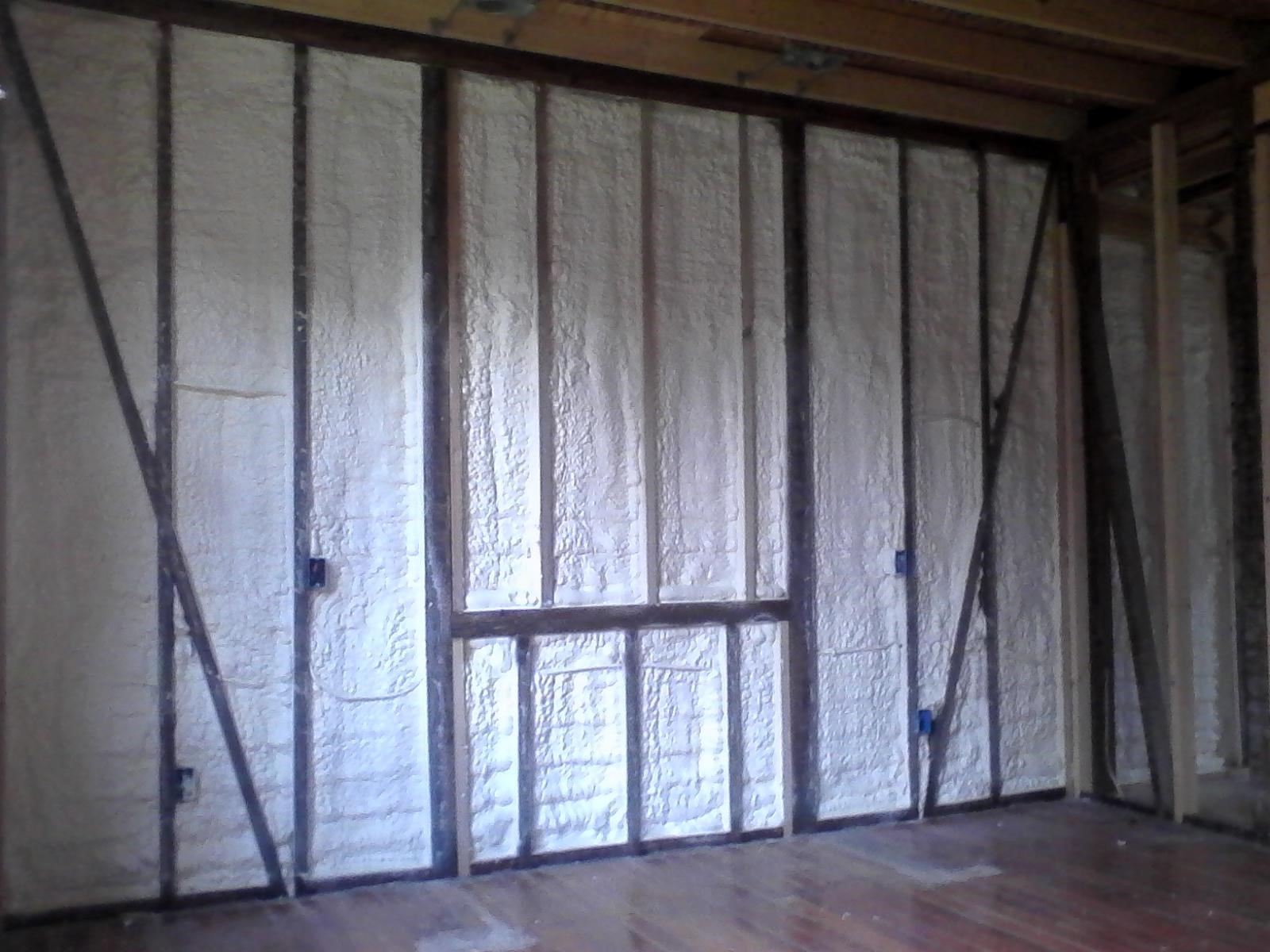 A room under construction with exposed wall studs showcases the meticulous work of an insulation company, with white spray foam insulation expertly filling the spaces between the studs. The wooden floor is bare, and no furniture or fixtures are present yet.
