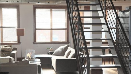 Modern living room with large windows, a staircase leading to an upper level, and a fireplace. The space features neutral-colored furniture and decor, ensuring comfort all year round thanks to the expertise of an insulation installer.
