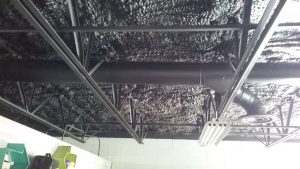 A ceiling with exposed black pipes and beams, black spray-foam insulation from a renowned insulation company in Baton Rouge, and a suspended fluorescent light fixture.