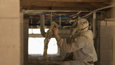 A person in protective gear applies foam insulation in a crawl space beneath a building, expertly provided by an insulation company in Baton Rouge.