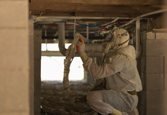 A worker in protective gear is crouched in a crawl space, inspecting and handling insulation material wrapped around pipes. The meticulous attention to detail suggests the expertise of an insulation contractor from a reputable insulation company.