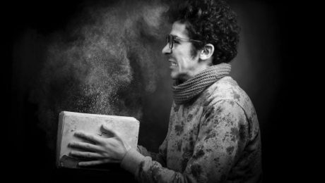 A person in glasses and a scarf, holding a book from which dust is emanating, is depicted in this black and white photo, reminiscent of an insulation contractor reading blueprints for an insulation company.