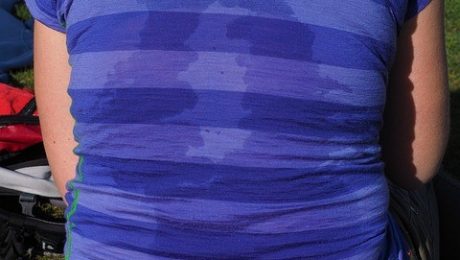 A person wearing a blue and purple striped shirt, with visible sweat marks, is shown from the back. They are seated on grass with a backpack nearby. Perhaps they could benefit from some advice from an insulation company Baton Rouge professionals to stay cooler next time.