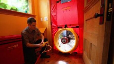 Blower-Door-Test A person kneels on a wooden floor, operating a device connected to a large fan installed in a doorway as part of a home energy audit. Their expertise in spray foam insulation ensures the home's energy efficiency is maximized.