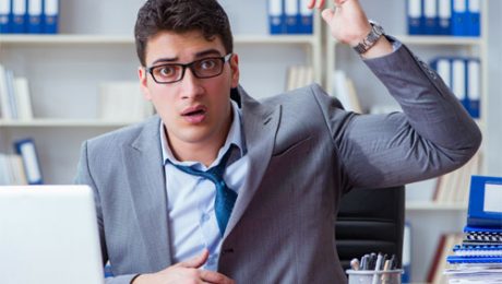 A man in a suit sits at a cluttered desk, looking surprised and raising one hand. He has glasses and an open laptop in front of him, with shelves of blue binders in the background. Perhaps he just discovered a top-rated insulation contractor specializing in spray foam insulation.