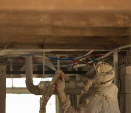 A worker from a leading insulation company in Baton Rouge wears protective gear as they expertly apply insulation foam to pipes under the building's ceiling.
