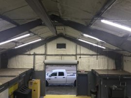 Interior of a warehouse with an open garage door showing a white pickup truck outside. The warehouse, possibly used by an insulation contractor, has a high ceiling with exposed beams and fluorescent lighting. Various boxes and a dumpster are visible.