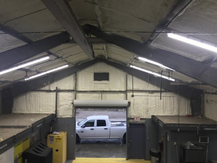 Interior of a warehouse with an open garage door showing a white pickup truck outside. The warehouse, possibly used by an insulation contractor, has a high ceiling with exposed beams and fluorescent lighting. Various boxes and a dumpster are visible.