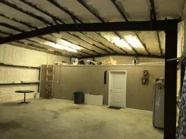 A spacious, insulated garage with metal beams, wall shelves holding various items, a ladder, a door, a mini fridge, and a small round table. The floor is concrete, and fluorescent lights hang from the ceiling—beautifully prepared by an insulation company for ultimate comfort.