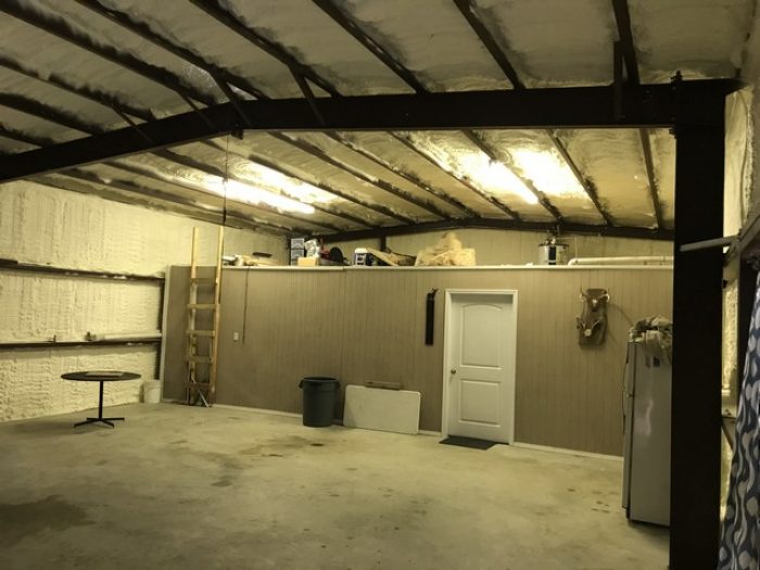 A spacious, insulated garage with metal beams, wall shelves holding various items, a ladder, a door, a mini fridge, and a small round table. The floor is concrete, and fluorescent lights hang from the ceiling—beautifully prepared by an insulation company for ultimate comfort.