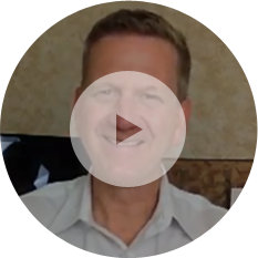 Thumbnail of a video featuring a man smiling, wearing a light-colored shirt, with a play button overlayed on his face. Plain background with a screen partially visible behind him, likely discussing the benefits of hiring an insulation contractor.