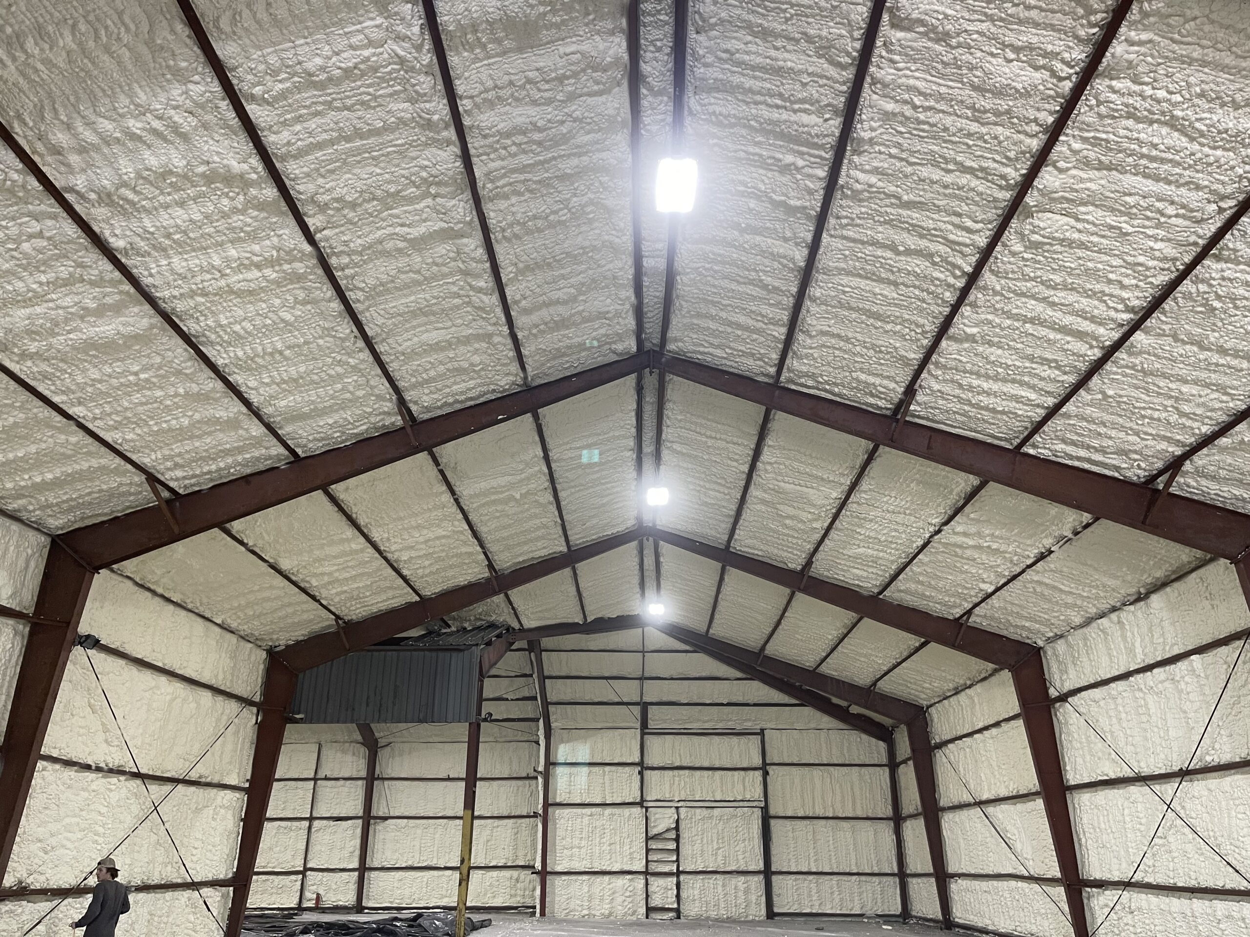 Interior of a large warehouse with metal beams and spray foam insulation covering the walls and ceiling; a person stands in the corner.